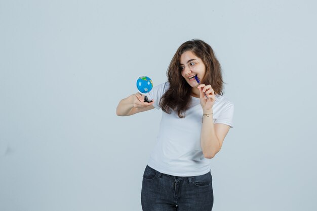 Young girl in t-shirt, jeans keeping pen on mouth while holding globe and looking happy , front view.