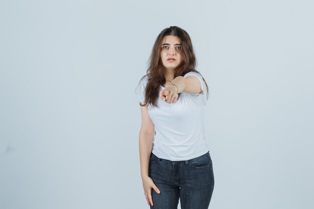 Молодая девушка в футболке, джинсах, указывающих на камеру и расстроенных, вид спереди.