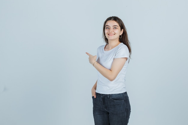 Young girl in t-shirt, jeans pointing to the left side and looking happy , front view.