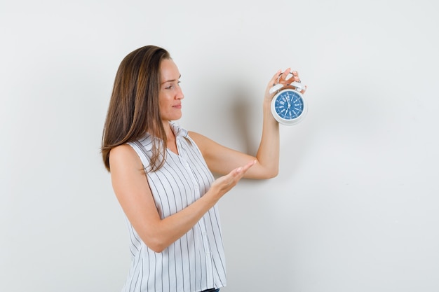 Young girl in t-shirt, jeans showing alarm clock and smiling , front view.