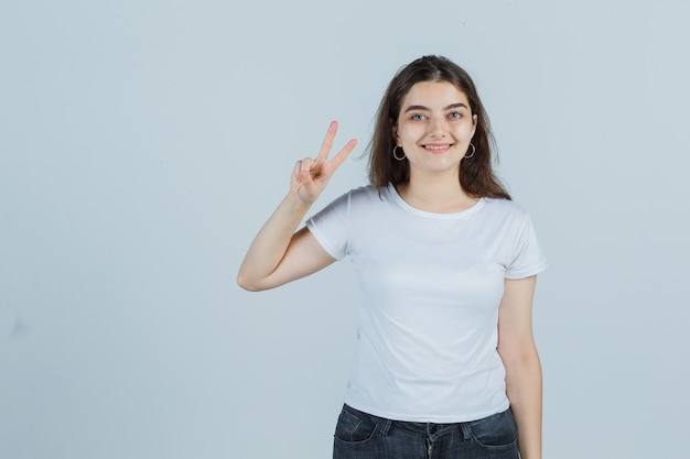 Молодая девушка в футболке, джинсах показывает знак победы и выглядит удачливым, вид спереди.