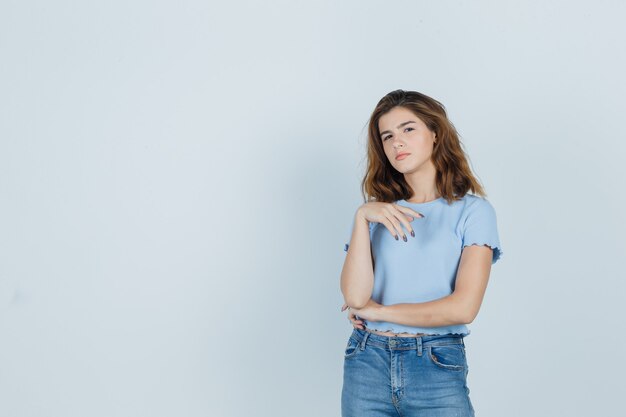 Молодая девушка в футболке, джинсах, стоящих в позе мышления и выглядящих разумно, вид спереди.