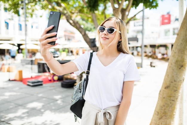 若い女の子は夏の街の通りで携帯電話で手から自分撮りを取ります。