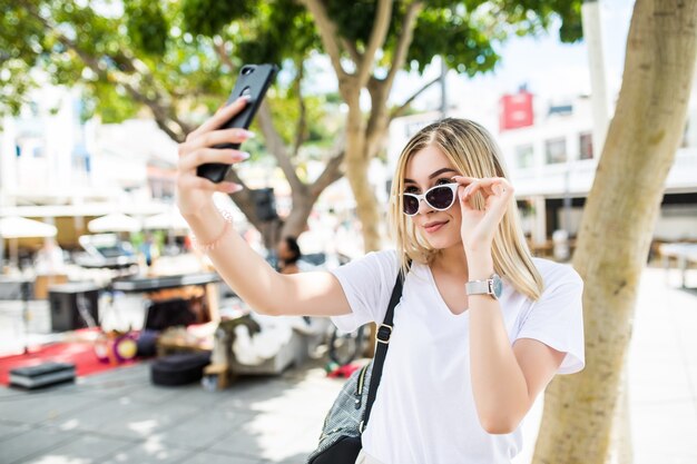 若い女の子は夏の街の通りで携帯電話で手から自分撮りを取ります。