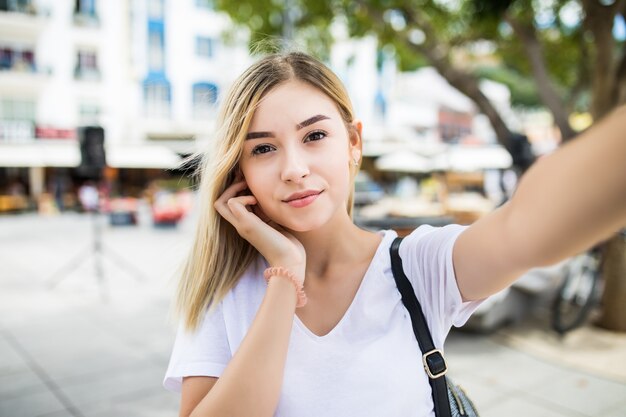 Молодая девушка принимает селфи из рук с телефоном на улице летнего города.
