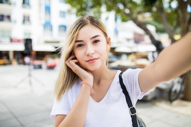 若い女の子は夏の街の通りで携帯電話で手から自分撮りを取ります。