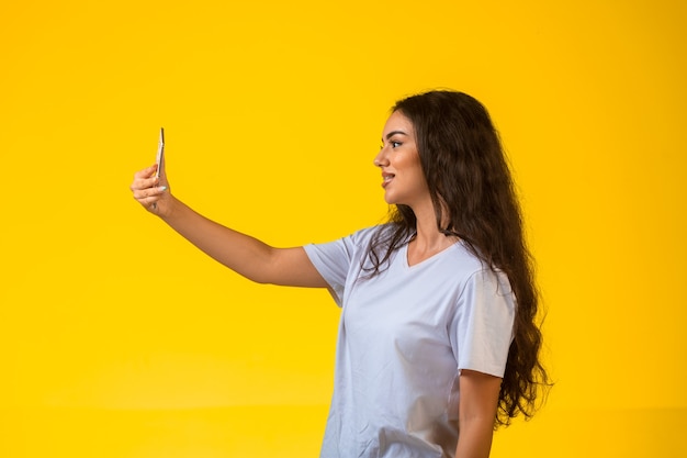 Молодая девушка принимает ее селфи на мобильном телефоне на желтом фоне и положительно улыбается, вид профиля.