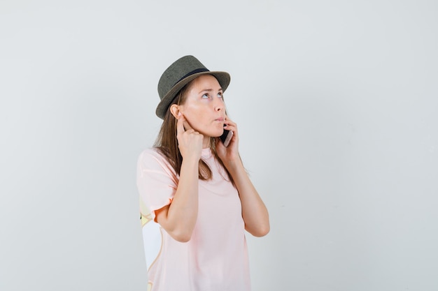 Young girl talking on mobile phone in pink t-shirt, hat and looking pensive. front view.
