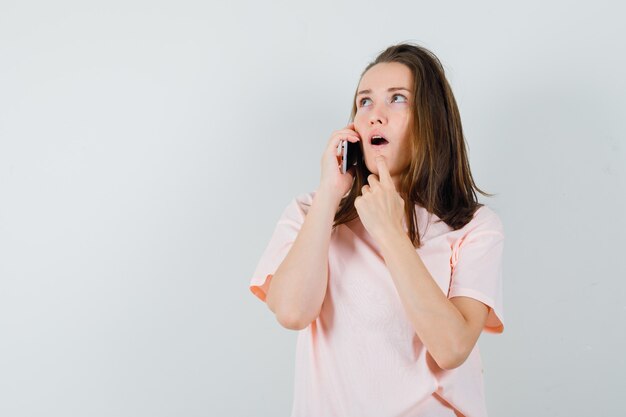 Молодая девушка разговаривает по мобильному телефону в розовой футболке и выглядит задумчиво