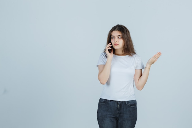 Young girl talking on mobile phone in t-shirt, jeans and looking serious. front view.