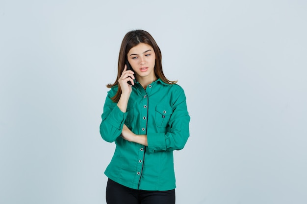 Young girl talking to phone, looking down in green blouse, black pants and looking focused , front view.