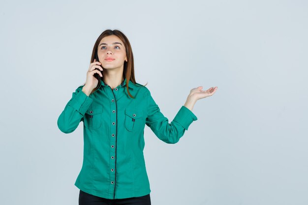 Young girl talking to phone, spreading palm aside in green blouse, black pants and looking happy , front view.
