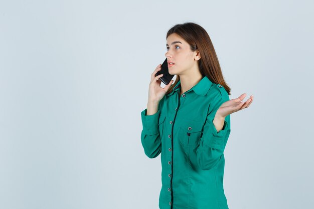 Young girl talking to phone, spreading palm out in green blouse, black pants and looking focused , front view.