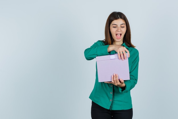 Young girl trying to open gift box in green blouse, black pants and looking excited. front view.