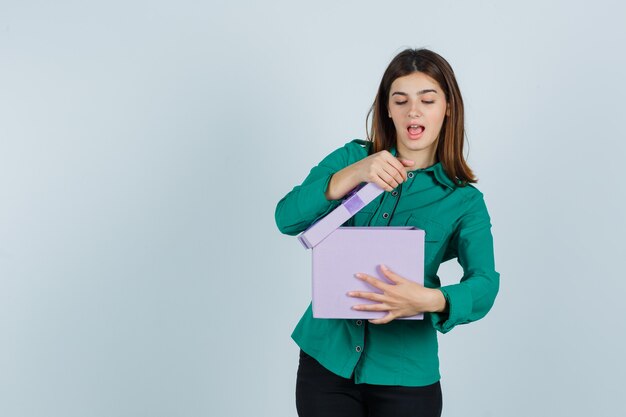 Young girl trying to open gift box in green blouse, black pants and looking excited. front view.