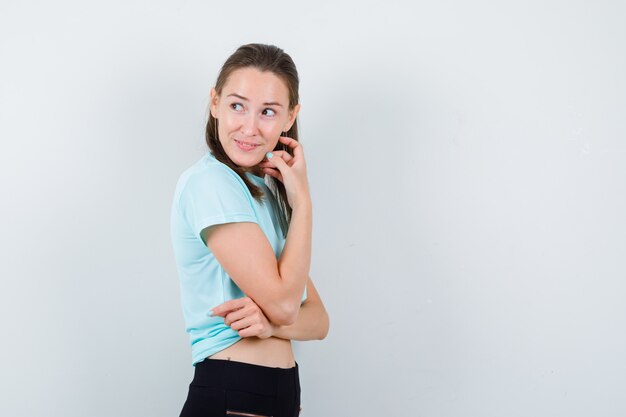 ターコイズ色のTシャツを着た少女、顔の近くに手があり、思慮深く見えるパンツ。