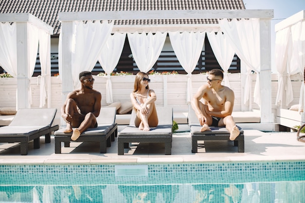 Young girl and two her multiracial male friends relaxing on a sunbeds near swimming pool. Girl wearing white swimwear and a sunglasses