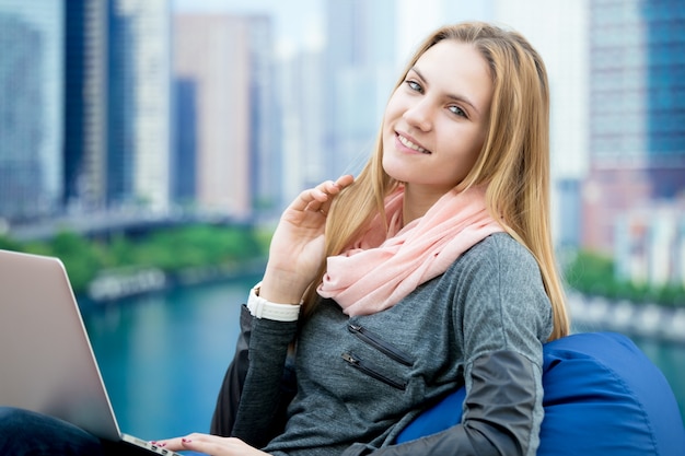 Free photo young girl using laptop in comfy armchair with big city on the background