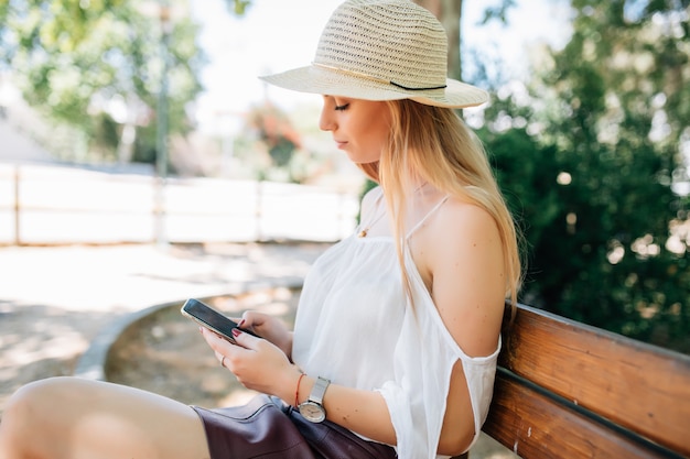 Ragazza giovane utilizzando uno smart phone e inviare messaggi di testo seduto su una panchina di un parco urbano