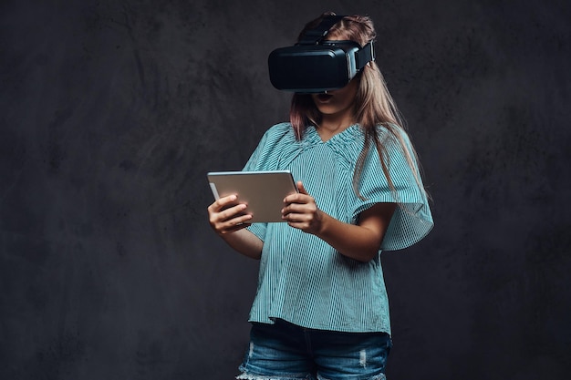 Young girl using virtual reality glasses and holds a tablet computer. Isolated on a dark textured background.