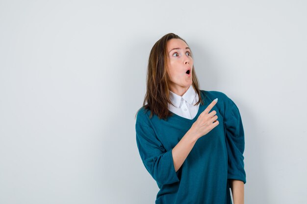 Young girl in v-neck sweater, shirt pointing up, opening mouth and looking shocked , front view.