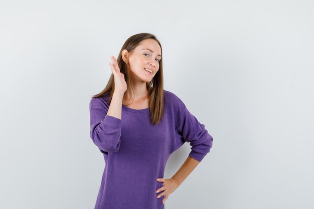 Молодая девушка в фиолетовой рубашке держит руку за ухом и смотрит любопытно, вид спереди.