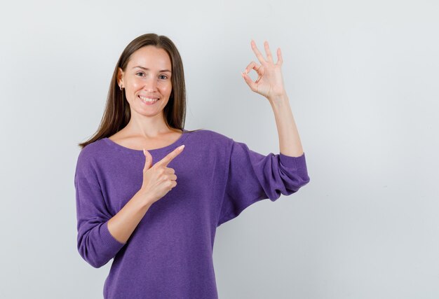 Young girl in violet shirt pointing at ok gesture and looking cheerful , front view.
