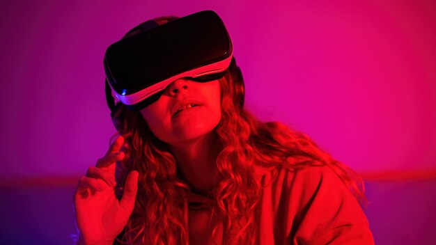 Young girl in virtual reality glasses with blue and red illumination in the room with rised up hand. Entertainment at home