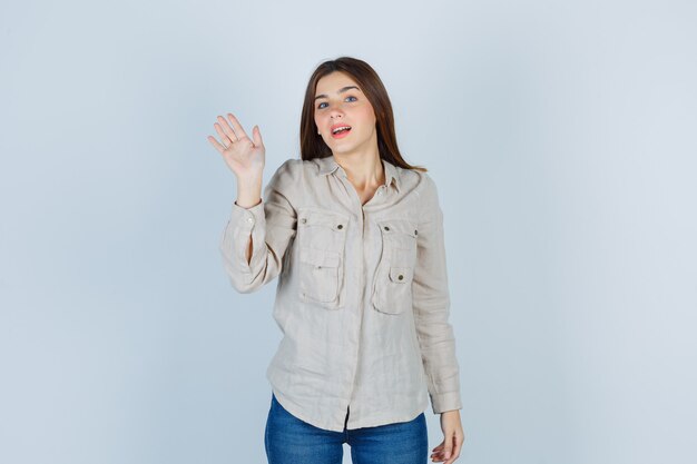 Young girl waving hand to greet someone in beige shirt, jeans and looking amiable. front view.