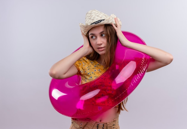 Young girl wearing hat and swim ring putting hands on head looking at left side on isolated white wall with copy space