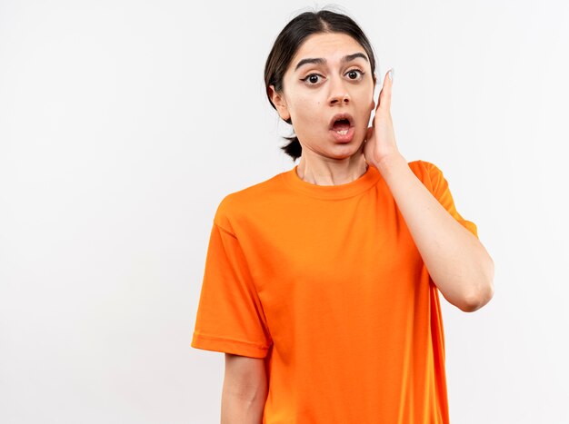 Молодая девушка в оранжевой футболке удивлена и смущена, стоя у белой стены