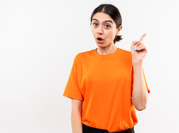 Молодая девушка в оранжевой футболке удивлена, показывая указательный палец, стоящий над белой стеной