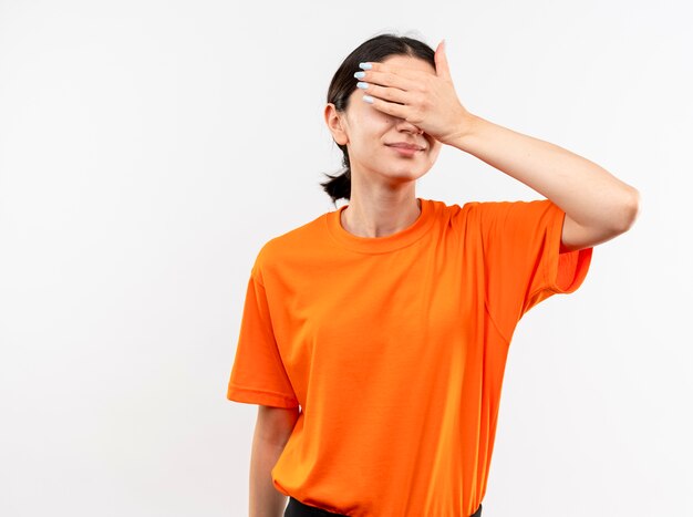 Young girl wearing orange t-shirt covering eyes with hand smiling standing over white wall
