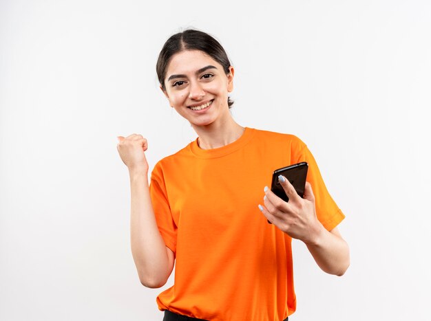 Young girl wearing orange t-shirt holding smartphone clenching fist happy and excited smiling cheerfully rejoicing her success standing over white wall