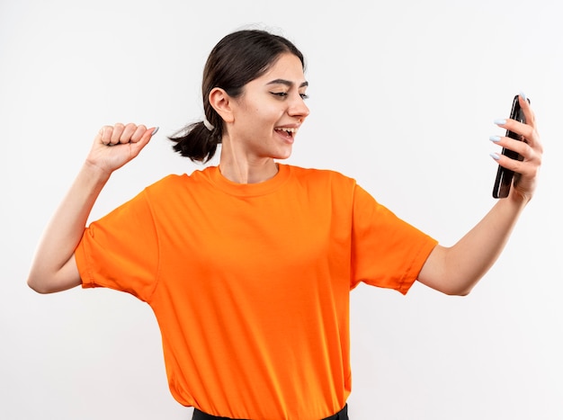 Young girl wearing orange t-shirt holding smartphone clenching fist happy and excited smiling cheerfully rejoicing her success standing over white wall