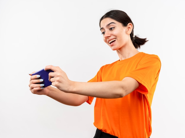 Giovane ragazza che indossa t-shirt arancione tenendo lo smartphone guardando sorridente felice e positivo in piedi sopra il muro bianco