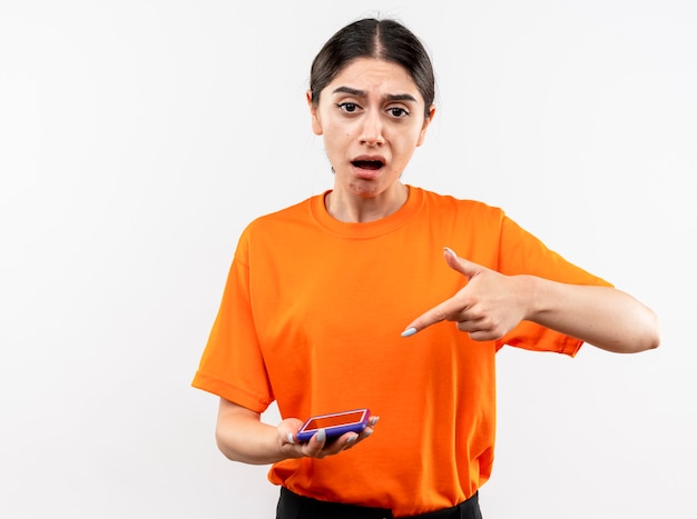 Молодая девушка в оранжевой футболке держит смартфон, указывая указательным пальцем на него, смущенная и недовольная, стоя у белой стены
