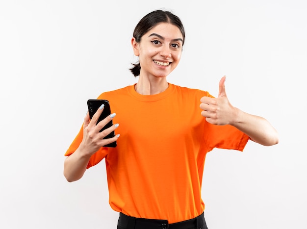 Young girl wearing orange t-shirt holding smartphone showing thumbs up smiling cheerfully standing over white wall