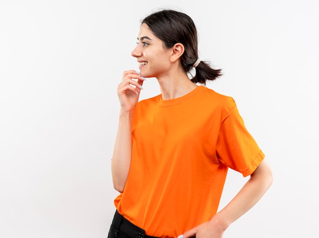 Young girl wearing orange t-shirt looking aside with hand on chin smiling with happy face standing over white wall