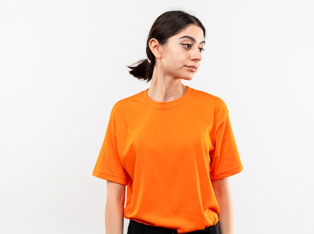 無料写真 白い壁の上に立っている悲しい表情で脇を見てオレンジ色のtシャツを着た少女