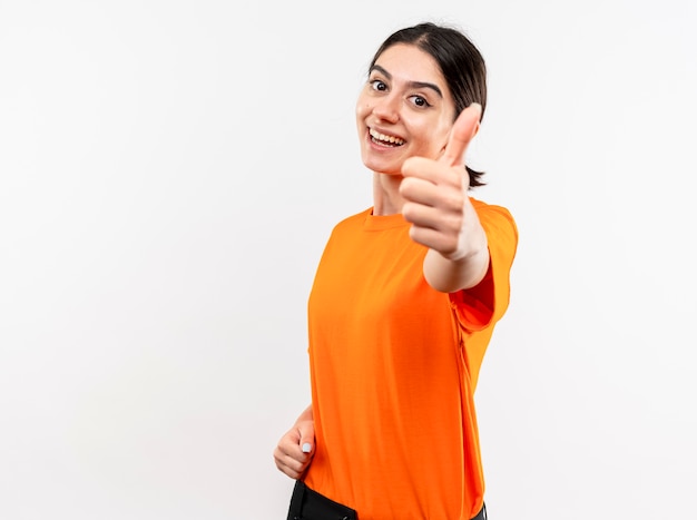 Young girl wearing orange t-shirt looking at camera smiling cheerfully showing thumbs up standing over white background