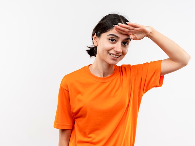 Молодая девушка в оранжевой футболке смотрит вдаль с рукой над головой, улыбаясь, стоя над белой стеной