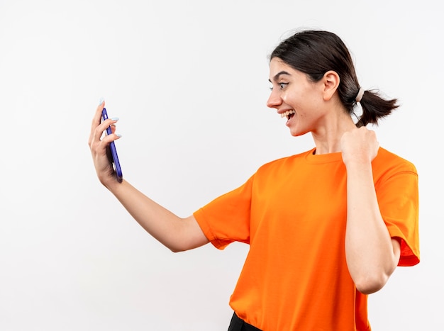 Молодая девушка в оранжевой футболке смотрит на экран своего смартфона, сжимая кулак, счастливая и позитивная улыбка, стоящая над белой стеной