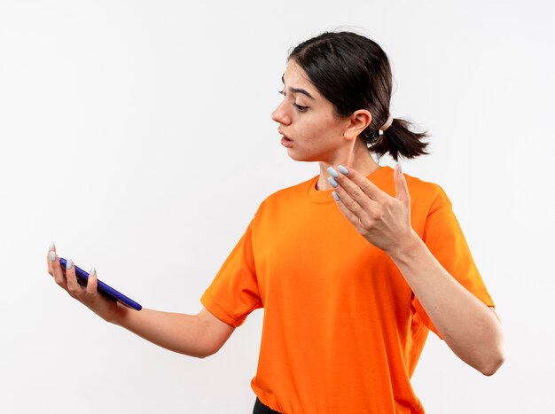 Young girl wearing orange t-shirt looking at her smartphone screen with arm out looking confused standing over white wall