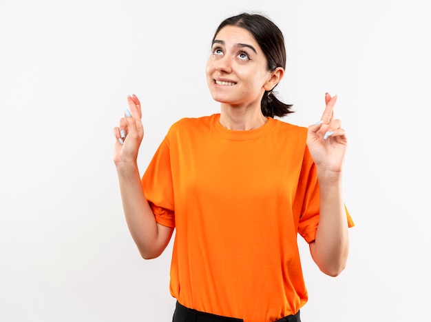 Young girl wearing orange t-shirt looking up biting lip making desirable wish crossing fingers standing over white wall