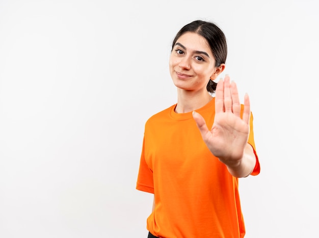 Молодая девушка в оранжевой футболке делает стоп-жест рукой с улыбкой на лице, стоящей над белой стеной