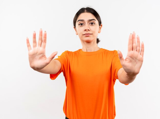 Молодая девушка в оранжевой футболке делает стоп-жест руками с серьезным лицом, стоящим над белой стеной