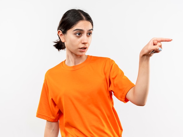 Young girl wearing orange t-shirt pointing with index finger at something being confused standing over white wall