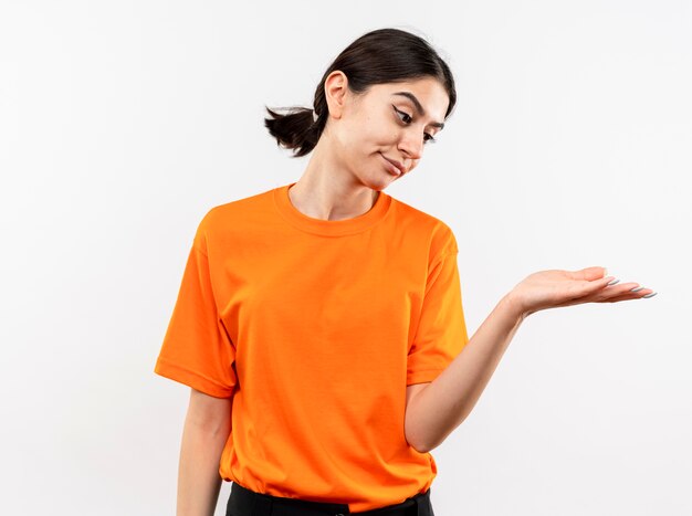 白い壁の上に立って不機嫌な手の腕で何かを提示オレンジ色のTシャツを着た少女
