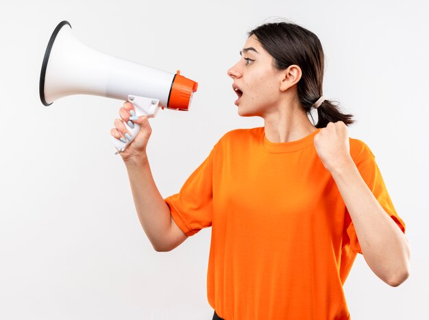 Young girl wearing orange t-shirt shouting to megaphone clenching fist emotional and excietd standing over white wall